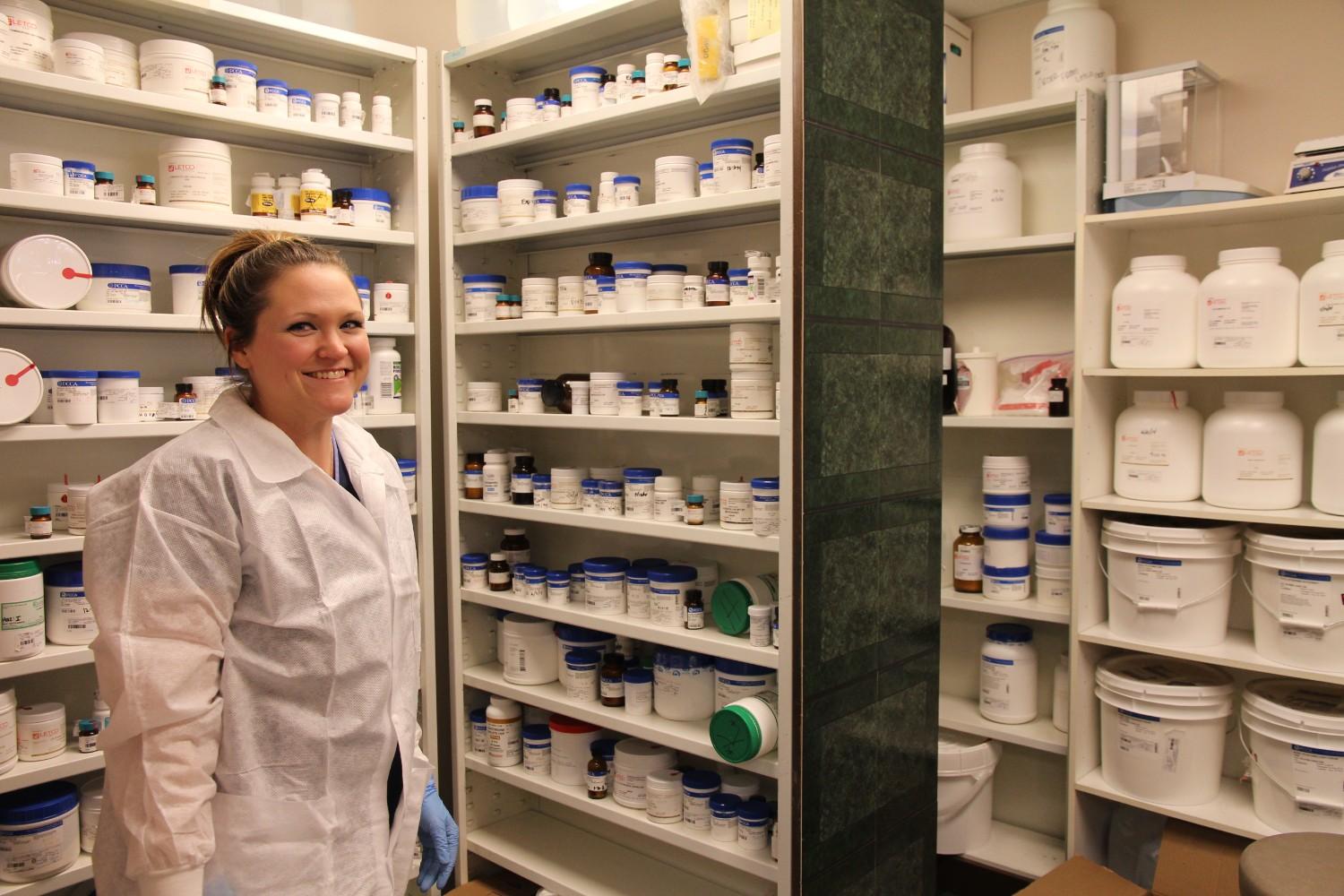 A smiling pharmacy technician in a white lab coat and blue gloves stands among floor-to-ceiling shelves stocked with medication 