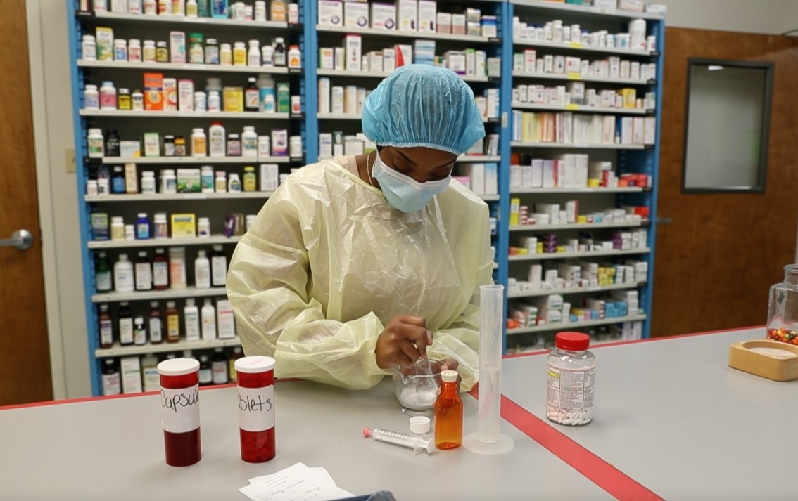 A pharmacy technician student in full protective gear measuring medications at a training lab