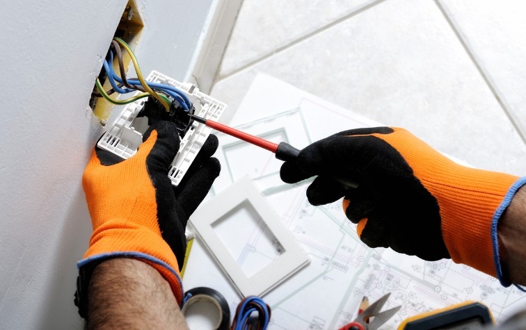 Close-up of an electrician's gloved hands using a screwdriver to wire an electrical outlet box in a wall