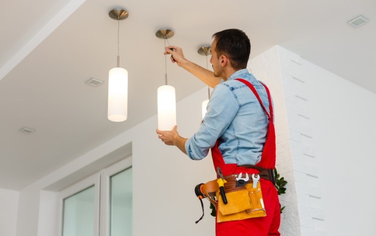 An electrician in red overalls and a tool belt installs pendant light fixtures on a ceiling inside a home
