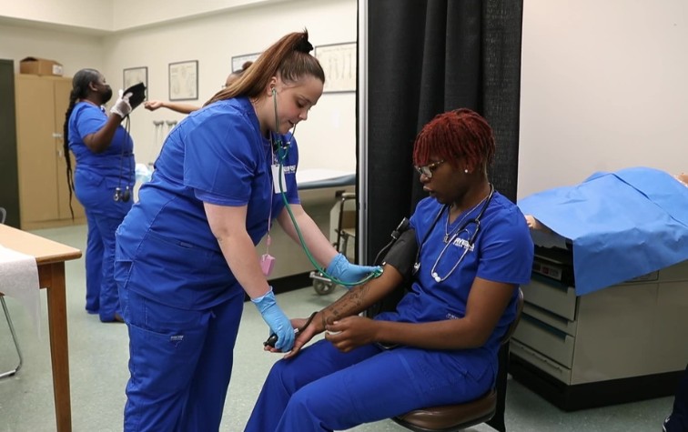 Medical assistant students in blue scrubs practicing taking blood pressure, with one student measuring another's arm using a cuff and stethoscope in a clinical training room.